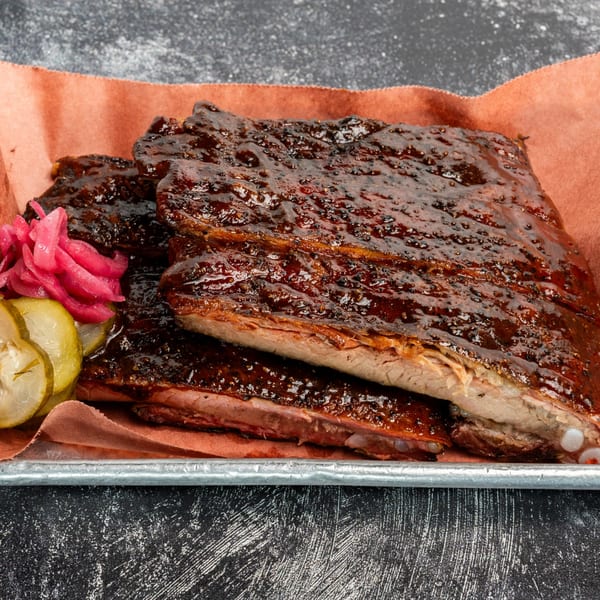 Creekside BBQ ribs, glistening with sauce, served with pickled onions and cucumber slices on a tray.