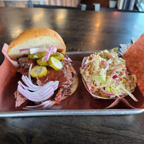 Creekside BBQ pulled pork sandwich with pickles, onions, and coleslaw on a metal tray.