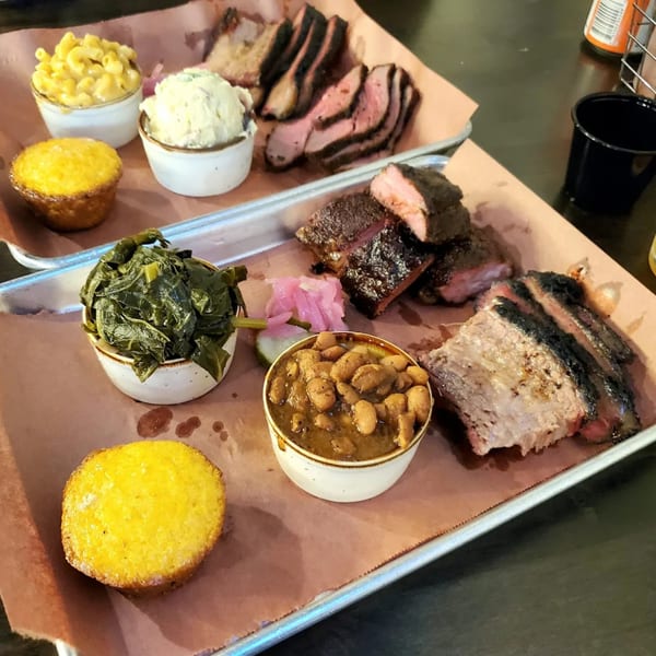 Two trays of Creekside BBQ with sliced brisket, ribs, mac and cheese, potato salad, collard greens, baked beans, and cornbread.